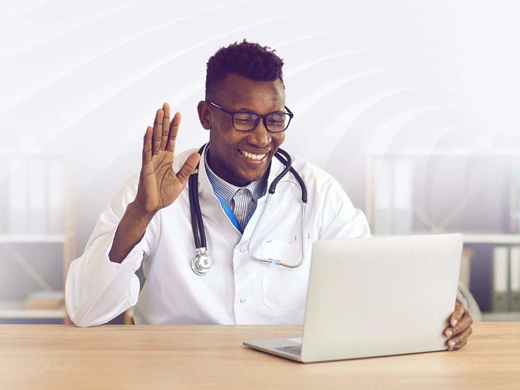 Doctor in a white coat with stethoscope waving hello at a laptop on a desk, as if greeting during a video call.